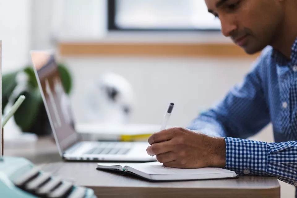 A man writing notes at a desk beside his laptop