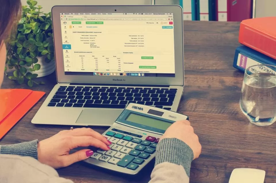 Hands using a calculator beside a laptop showing a spreadsheet